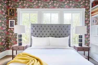 Eclectic bedroom with floral wallpaper, gray upholstered bedroom, yellow blanket, nightstands, lamps.